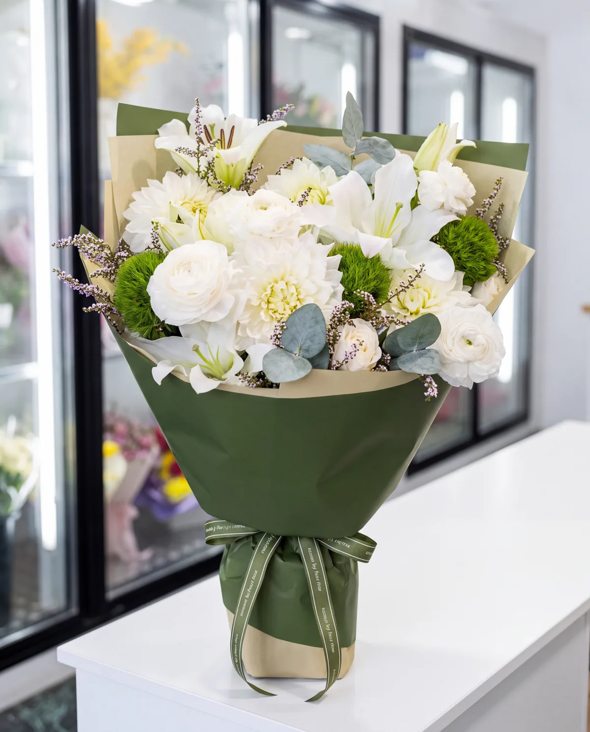 White ranunculus, dahlias and lilies with green button chrysanthemums and eucalyptus, in deep green wrap, on a white bench